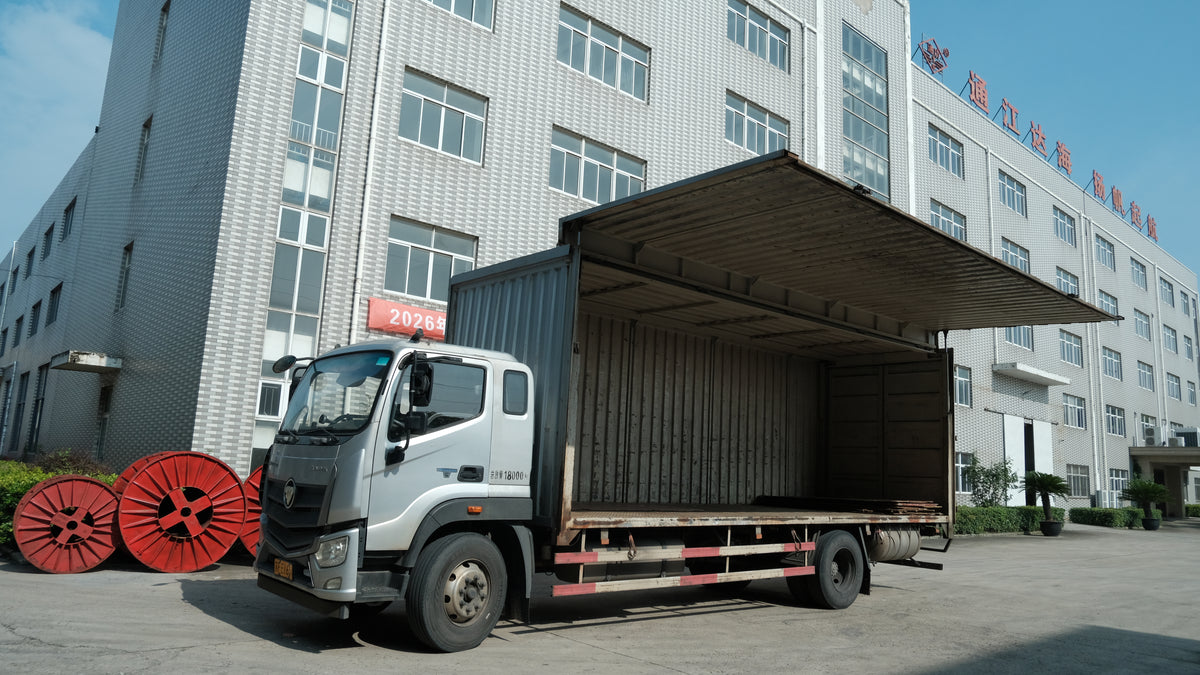 A white box truck with its cargo door open, parked in front of a large gray industrial building. Red spool-like objects are stacked on the left, and the truck is photographed outdoors under daylight.