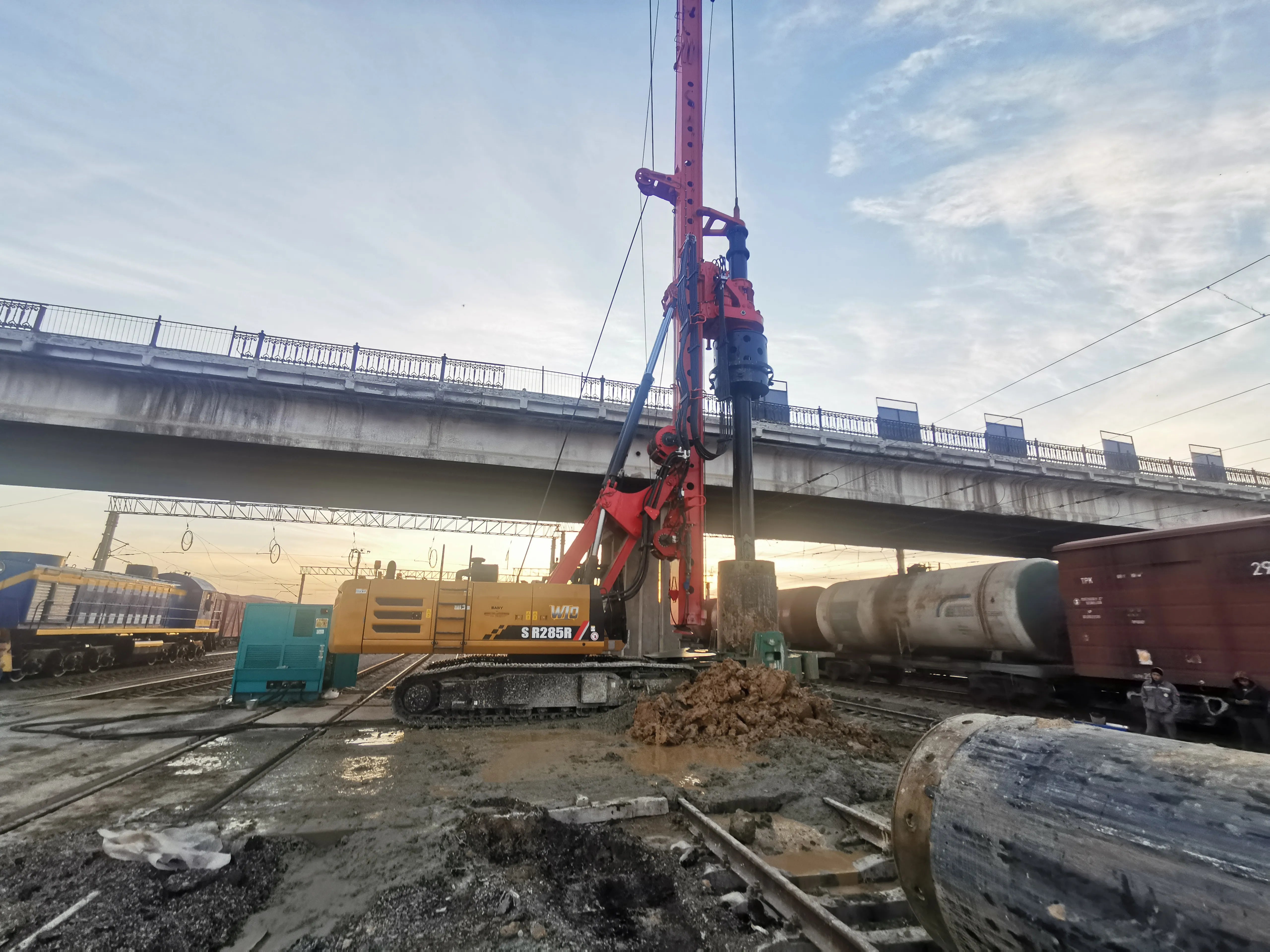 A red and black track-compacting crawler machine operating on railway tracks under an elevated bridge, with freight trains and industrial equipment in the background.