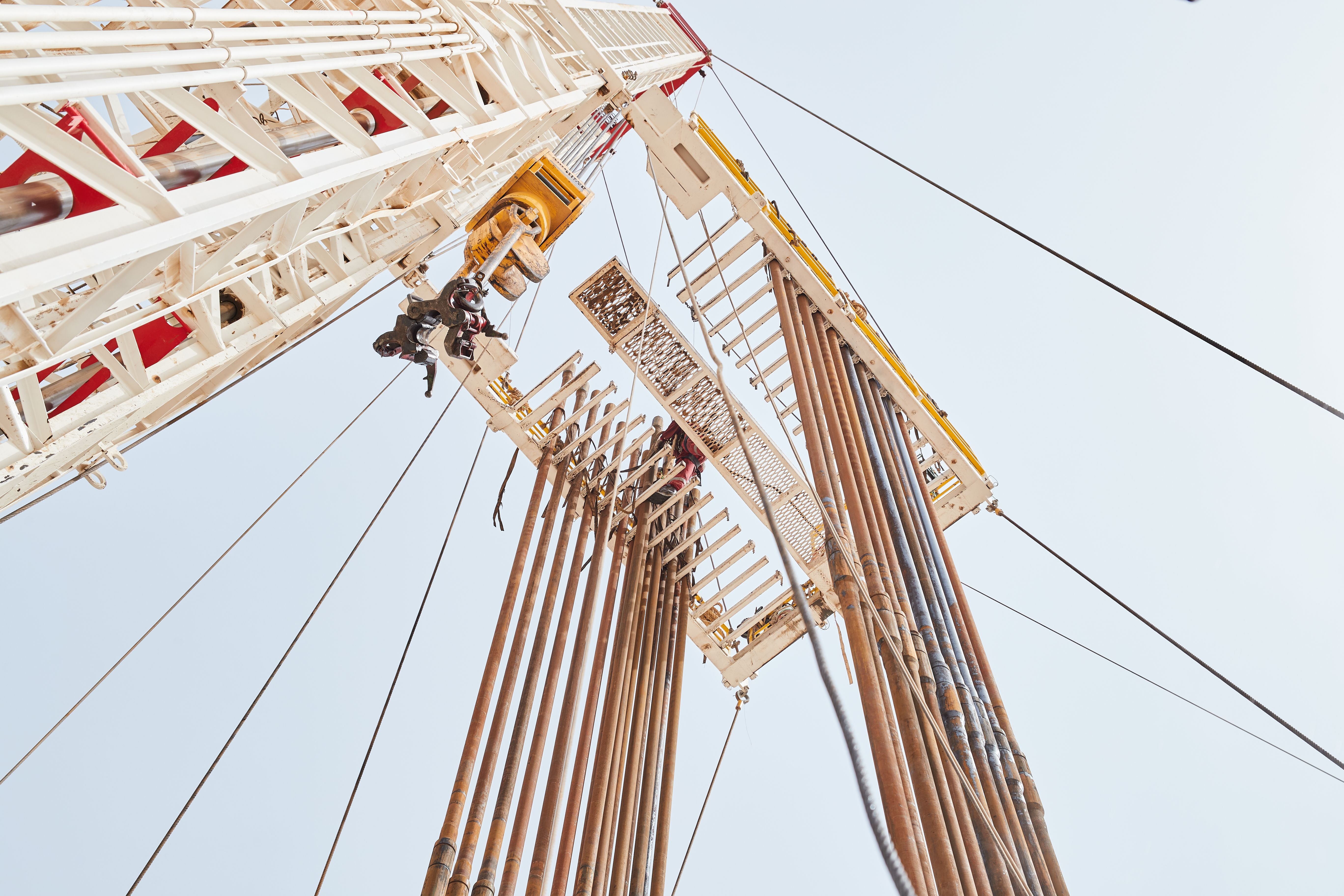 An oil rig drilling platform structure photographed from a low angle against a pale sky, with workers visible on the rig and multiple steel pipes and cables extending upward.
