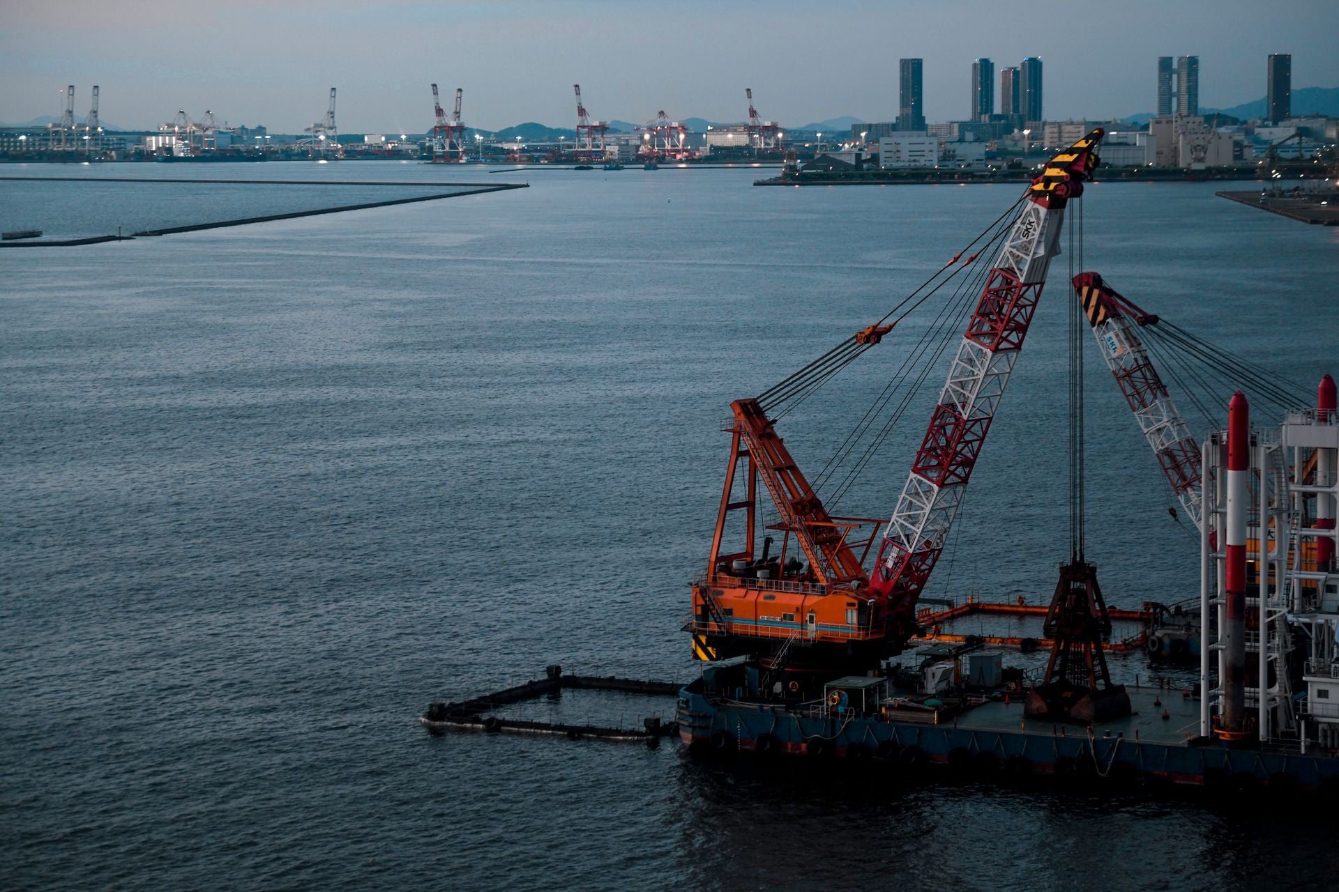 A large orange and white ship-mounted crane on a barge in a harbor, with water, distant industrial buildings, and cranes visible under a dim sky.