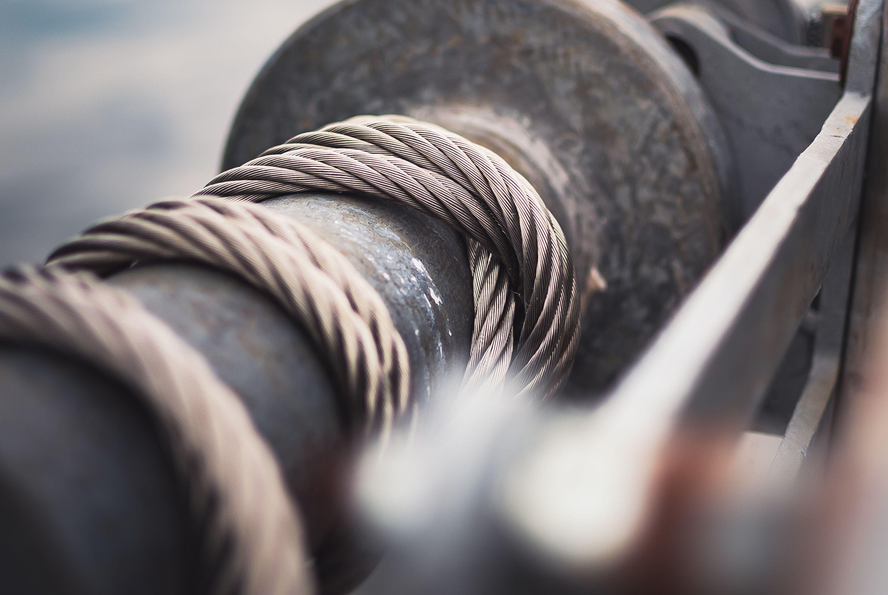 Close-up of a thick steel cable wrapped around a dark metal spool or winch component, with shallow depth of field and blurred foreground and background details.