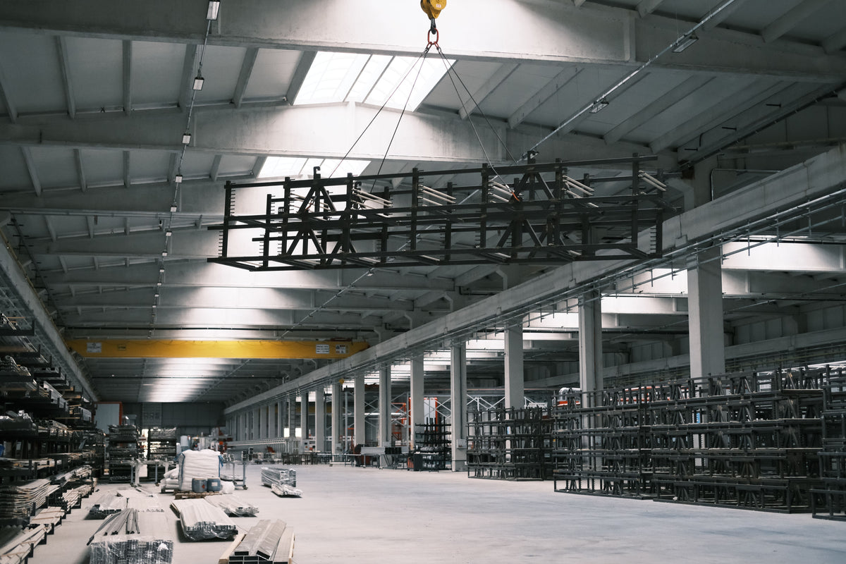 A large black metal warehouse shelving or rack frame suspended by a crane inside a high-ceilinged industrial warehouse with concrete floors, support columns, shelving units, and skylights.