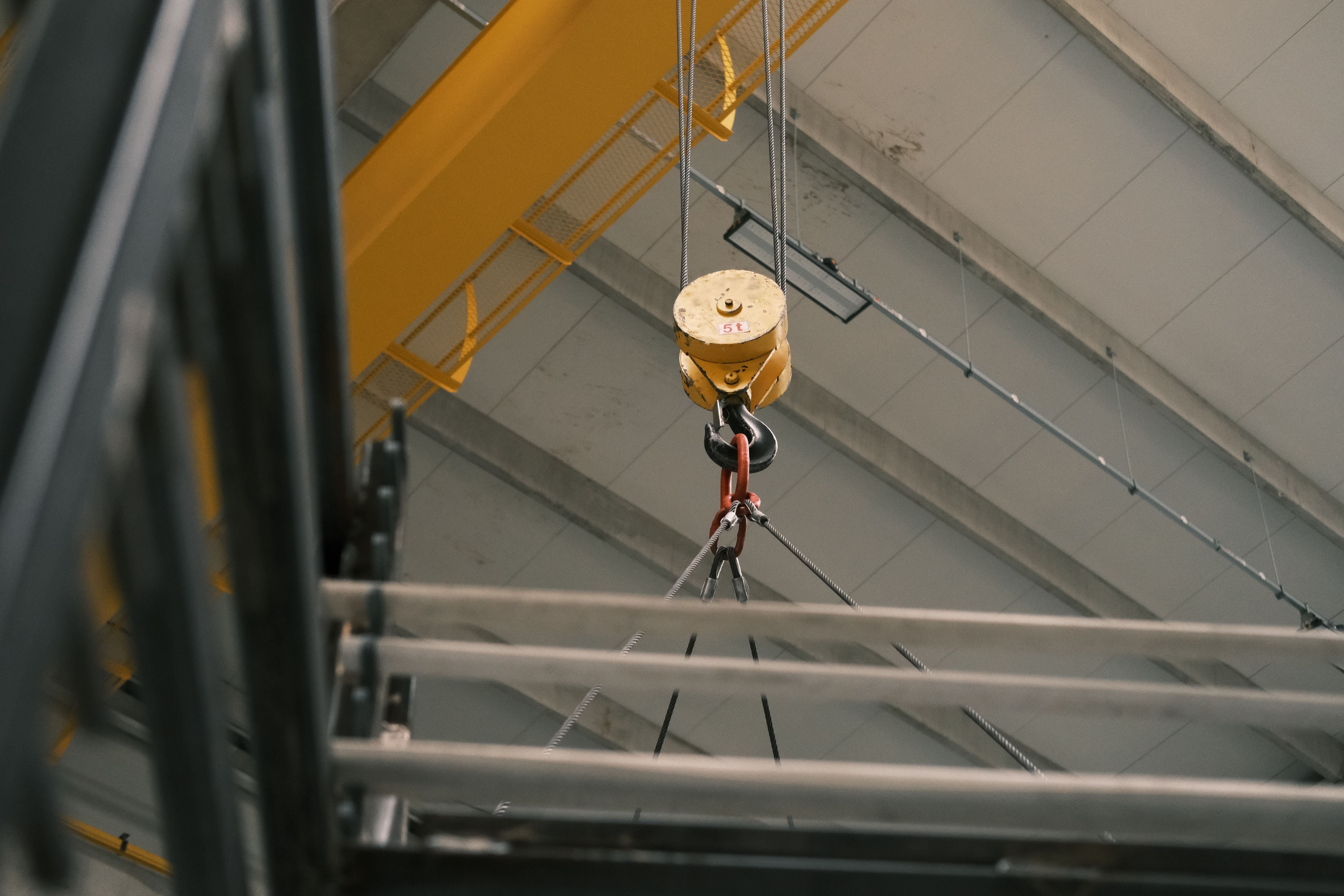 A close-up view of a yellow industrial overhead crane hook suspended by cables from a metal ceiling structure inside a warehouse or factory.