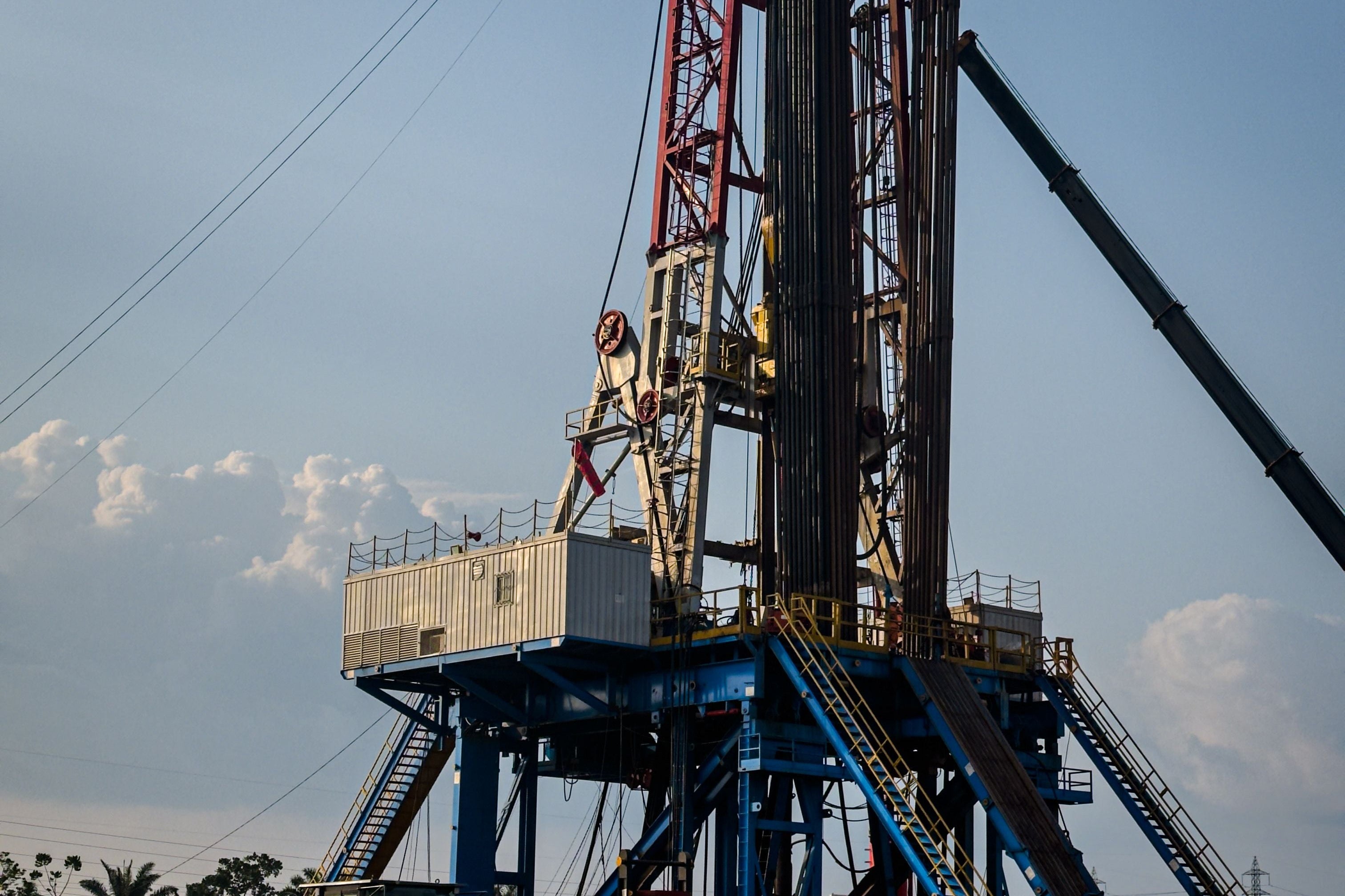 A large blue and red drilling rig stands on a paved outdoor lot under a blue sky, with a yellow construction vehicle parked nearby and a small worker visible near the base.