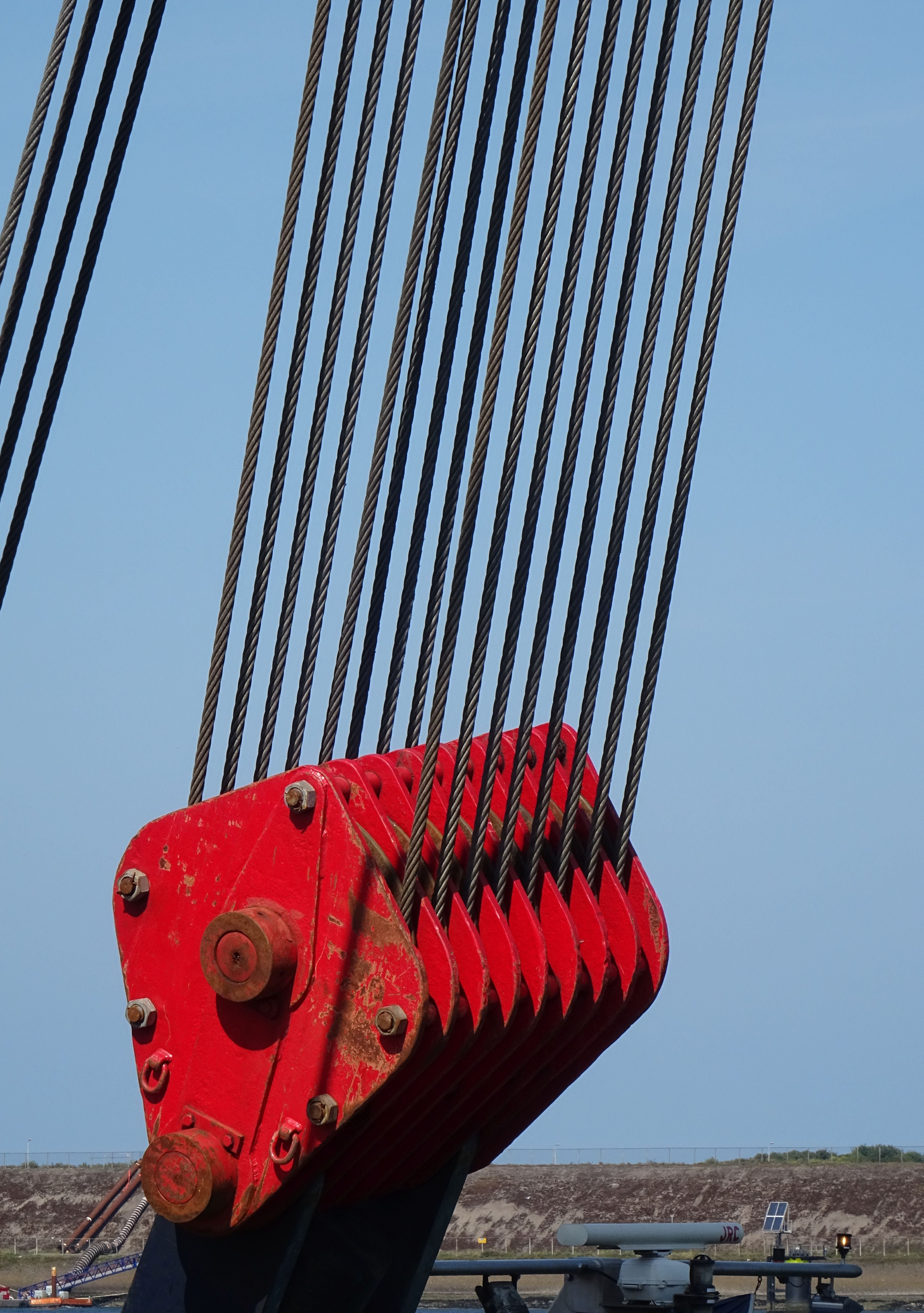 A close-up of a red heavy-duty steel cable hook attachment with multiple thick black steel cables, set against a clear blue sky with a distant construction site.