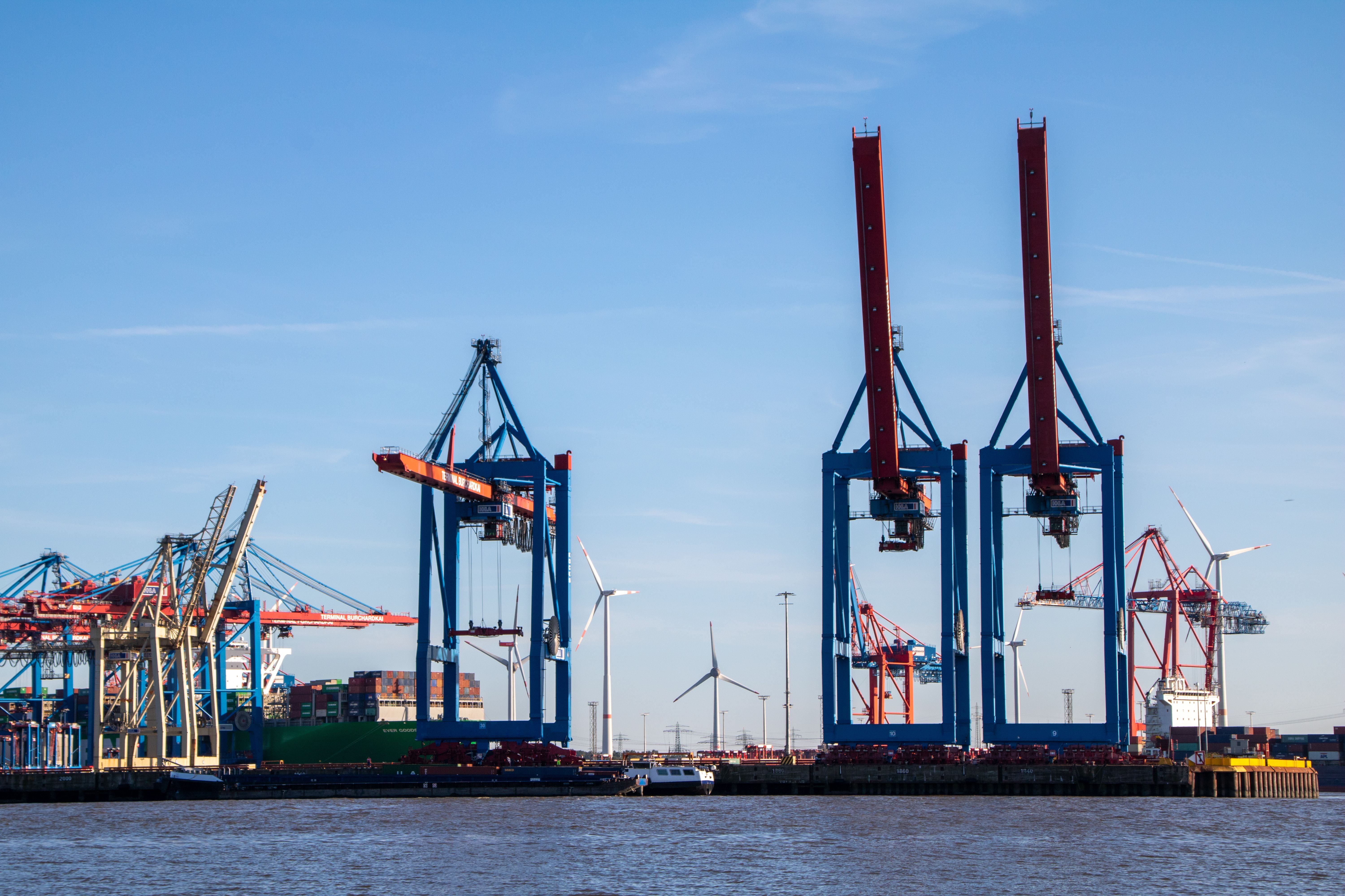 A wide outdoor harbor scene with large blue and red gantry cranes, wind turbines, and calm water under a clear blue sky.