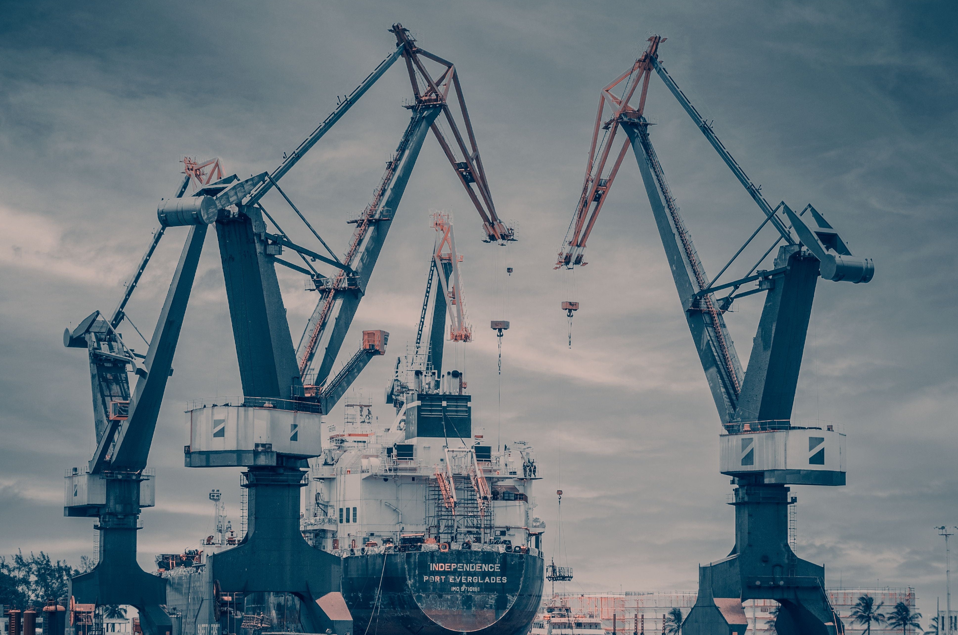A moody, desaturated photograph of a shipyard with two large cranes framing a large cargo ship on the water under a cloudy blue sky.