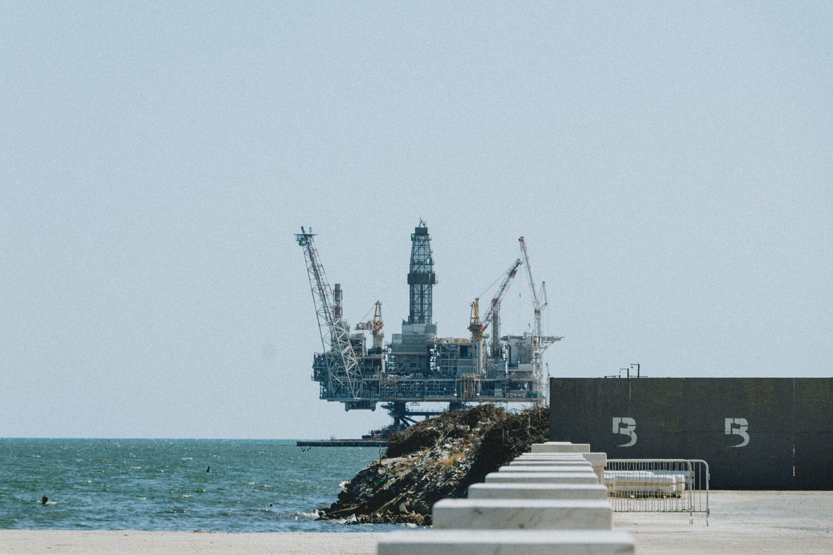 An offshore oil rig stands in the water beside a rocky pier and a concrete wall marked with the number 5, under a pale blue sky.