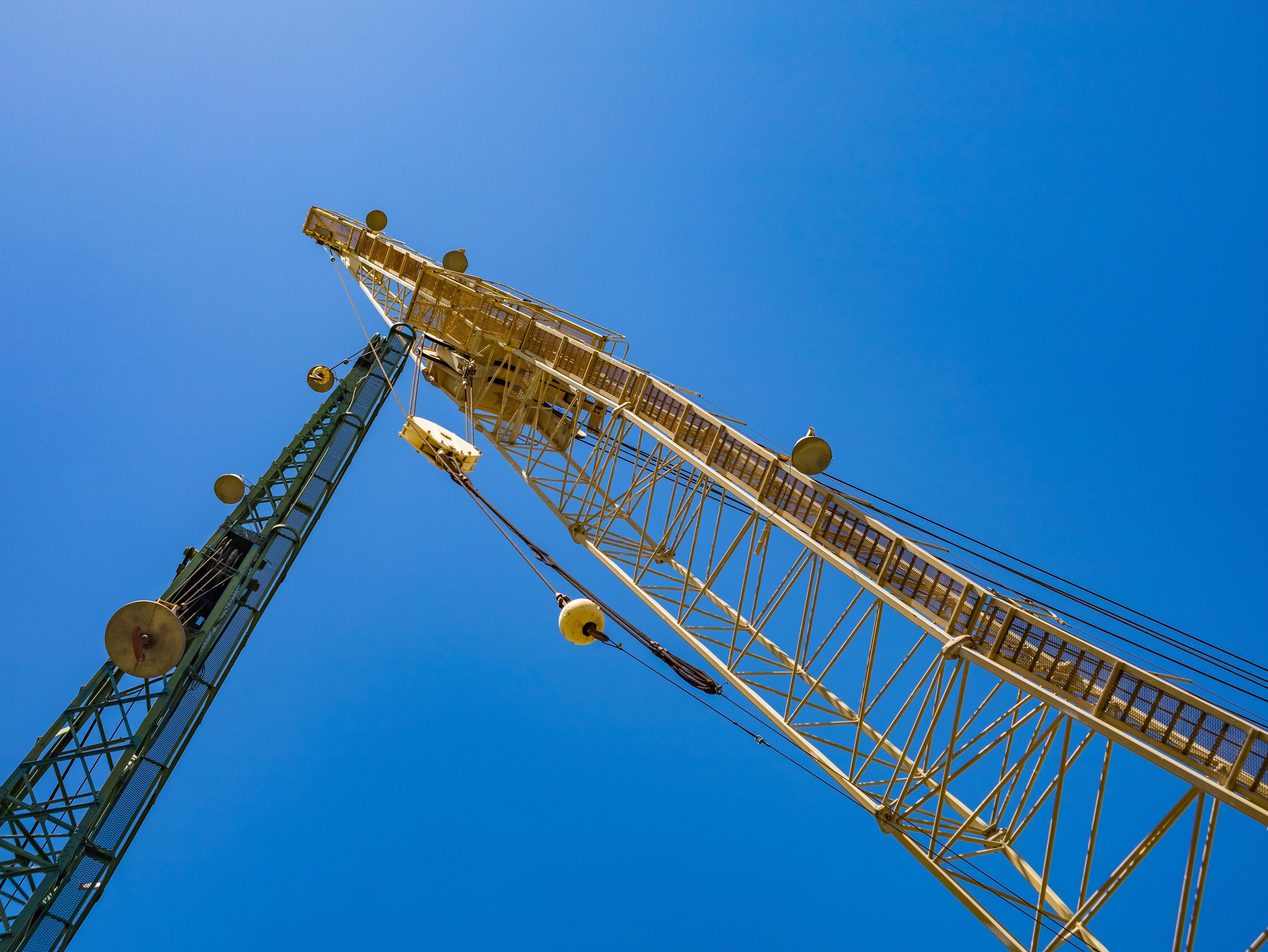 A low-angle view looking up at the yellow lattice boom of a large construction crane against a clear blue sky, with part of the green counterweight arm visible on the left.