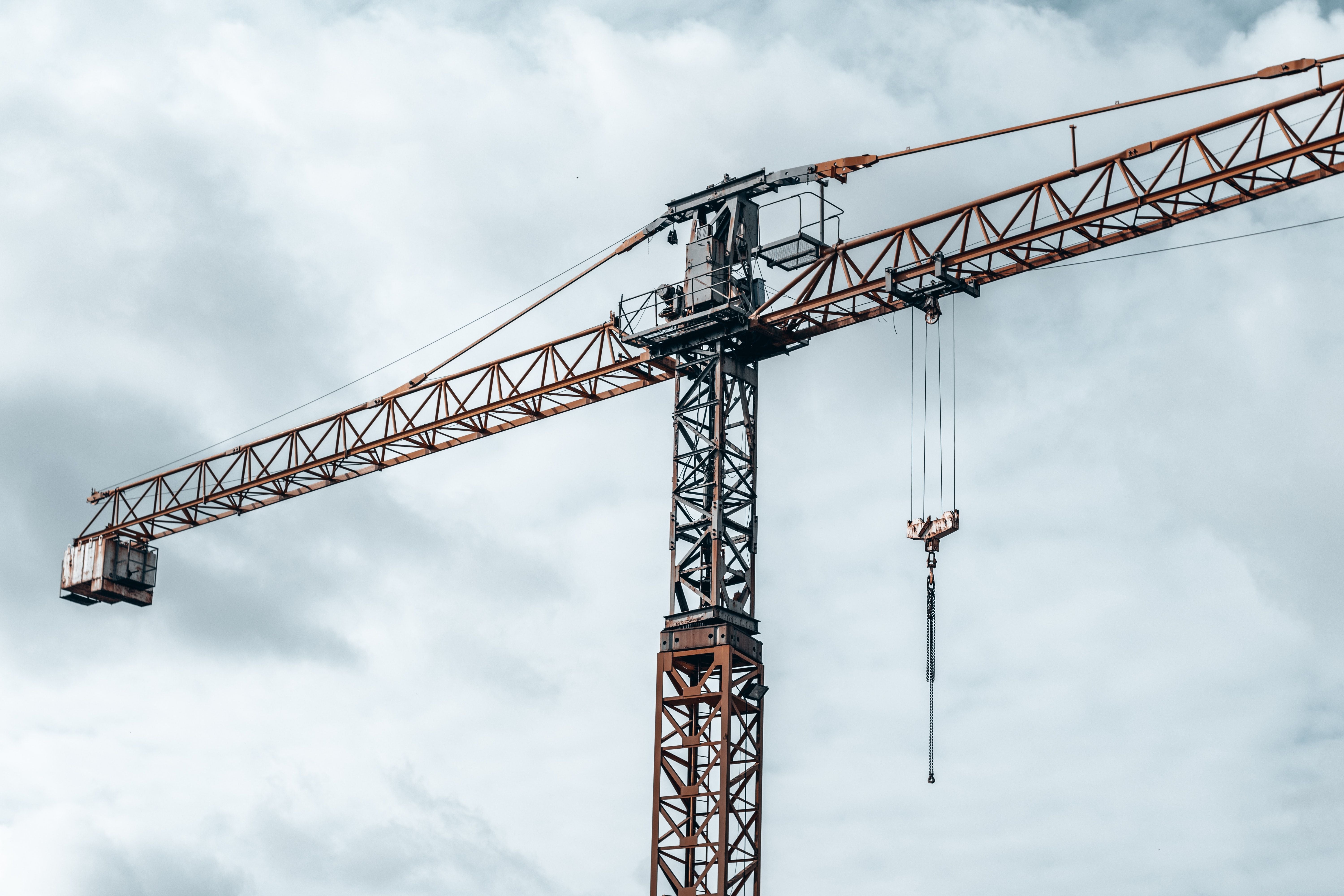 A large tower crane with a rusted orange arm and dark steel lattice structure, suspended against a cloudy white and gray sky.