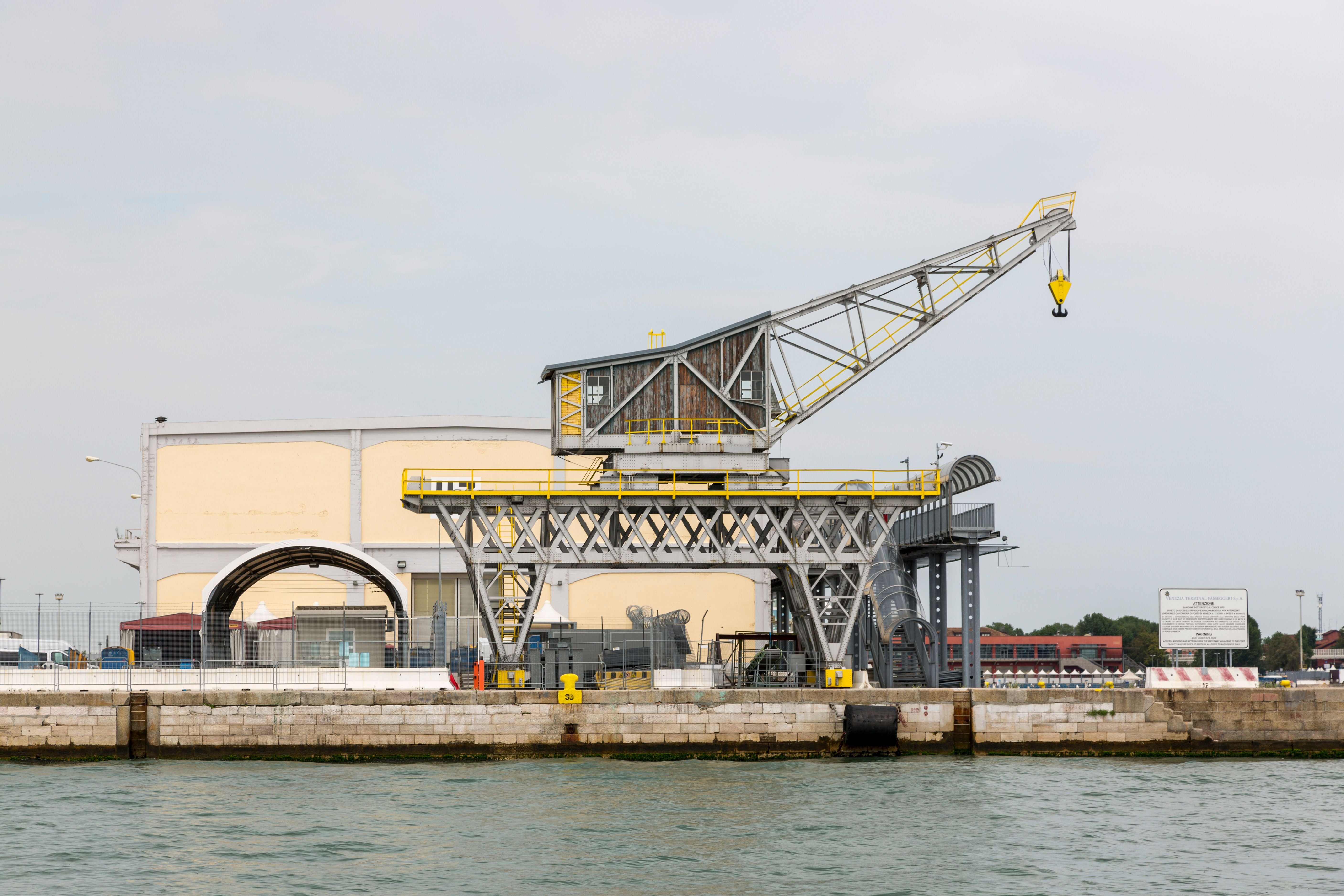 A large industrial gantry crane with yellow accents stands beside a waterfront pier, with a pale building and water in the background under an overcast sky.