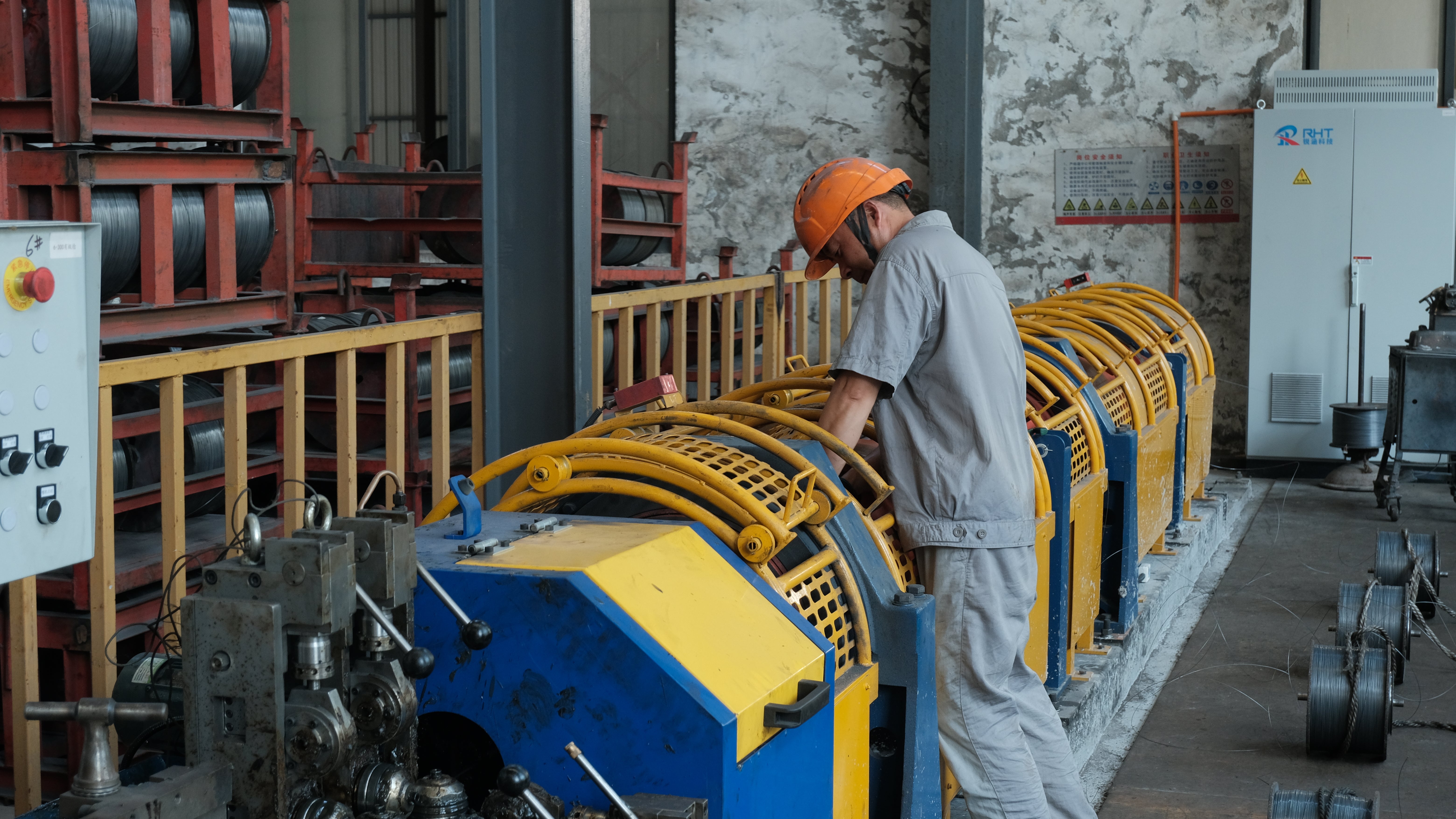 A worker in a gray uniform and orange hard hat operates a large yellow and blue industrial machine inside a factory or workshop with concrete walls and metal equipment.